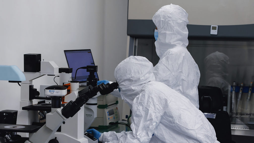 Two Immunocine scientists in full protective cleanroom suits working at a microscope.