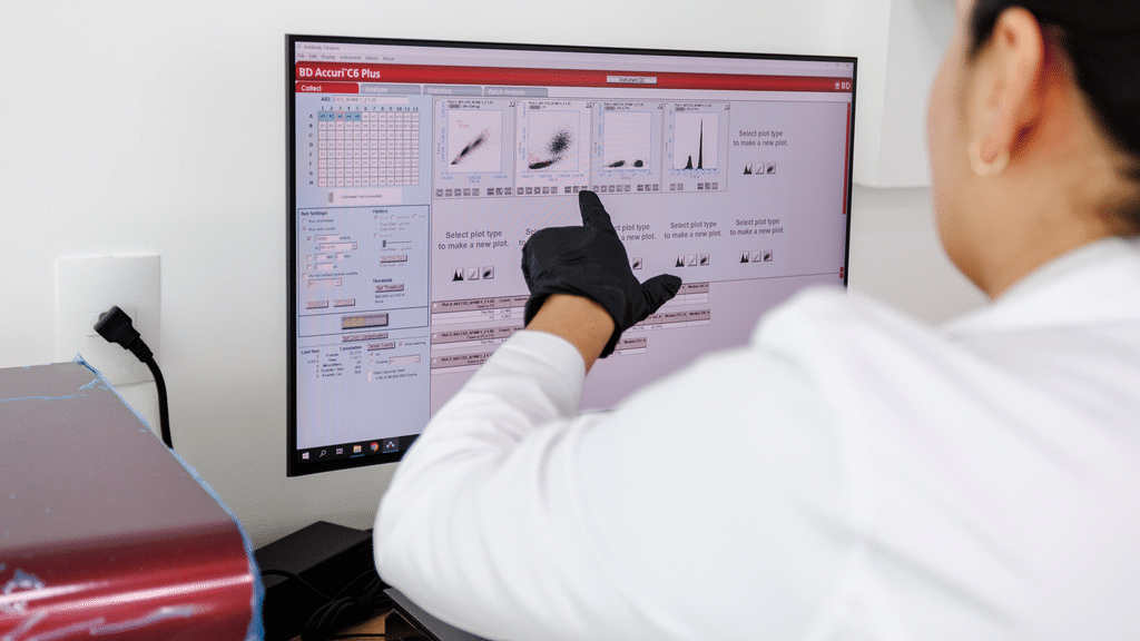 Immunocine lab scientist in gloves pointing at flow cytometry data on a computer monitor.